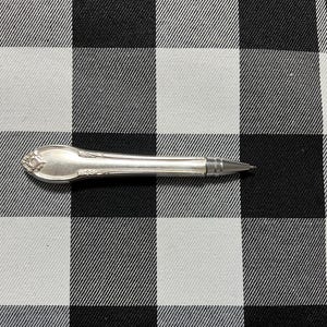 May include: A silver pen with a spoon-shaped end and a pointed tip. The pen has a decorative floral design on the spoon end and is lying on a black and white checkered surface.