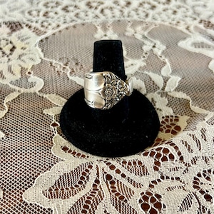 May include: A silver-toned ring crafted from a repurposed spoon, featuring an intricate embossed floral pattern on the front surface. The metal band displays a polished finish with detailed engravings of petals and leaves across the center.