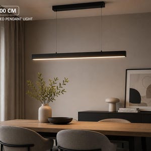 Può includere: Una lampada a sospensione LED lineare da 100 cm con finitura nero opaco, sospesa tramite due fili sottili da un supporto rettangolare a soffitto. L'apparecchio emette una luce calda rivolta verso il basso lungo la sottile barra orizzontale.
