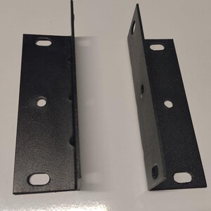 Puede incluir: Dos soportes metálicos negros en forma de L, cada uno con un ángulo recto. Cada soporte tiene un lado vertical plano con dos orificios de montaje ovalados y un orificio circular en el centro, diseñados para una fijación y soporte seguros.