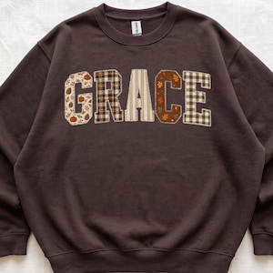 May include: A dark brown crewneck sweatshirt featuring the word GRACE in large, block-style letters across the chest. Each letter is filled with a different fabric pattern, including pumpkins, plaid, stripes, autumn leaves, and a green gingham check.