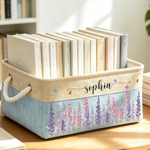 May include: A rectangular fabric storage bin with a beige upper section and a light blue lower section featuring a floral print of purple and pink lupine flowers. The name Sophia is printed in black script, accented by small colorful butterflies.