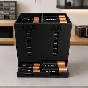 May include: A black plastic battery organizer with two vertical compartments labeled AAA and AA. The unit features top-loading slots for battery storage and two front-facing dispensing trays at the bottom, currently holding several black and copper Duracell batteries.
