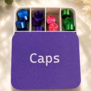 Könnte beinhalten: Eine violette Aufbewahrungsbox mit dem Wort Caps in weißem Aufdruck auf der Vorderseite. Die Box verfügt über ein offenes oberes Fach, das in vier Abschnitte unterteilt ist und kleine, metallische zylindrische Kappen in Blau, Lila, Pink und Grün enthält.
