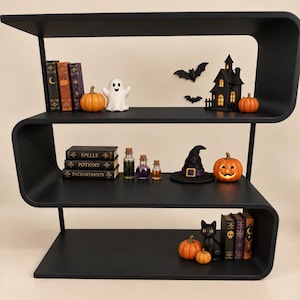 May include: A black S-shaped three-tier shelf decorated with Halloween items including miniature pumpkins, a ghost figurine, a haunted house, a witch hat, a black cat, and books labeled Spells, Potions, and Enchantments with glass vials.