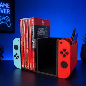 Puede incluir: Un organizador de juegos de plástico negro con forma de mando de consola, con el lado izquierdo azul claro y el derecho rojo. Contiene varias cajas de juegos y cuenta con un portalápices integrado en el lado derecho, mostrando una caja roja.