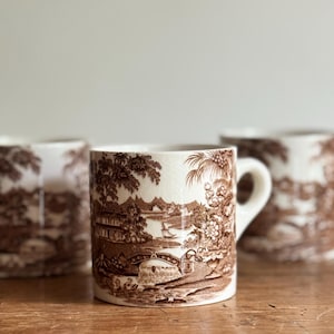 May include: Set of four ceramic mugs with a brown transferware design. Each mug features a detailed landscape scene with bridges, buildings, and trees. The mugs have a classic shape with a handle and a cream-colored background.