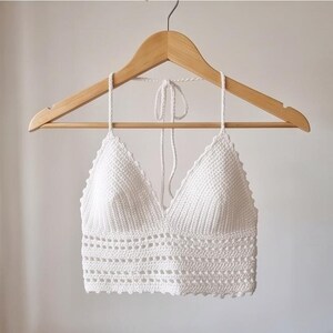 Pode incluir: Top de crochê branco com decote em V, exibido em um cabide de madeira. O padrão de crochê apresenta um ponto texturizado na parte superior e um design de trabalho aberto decorativo na borda inferior.
