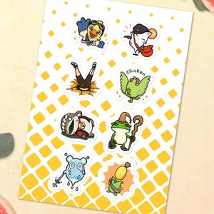 Könnte beinhalten: Ein Bogen mit Cartoon-Aufklebern mit verschiedenen skurrilen Charakteren. Die Aufkleber zeigen eine Ente mit Pilotenmütze, eine Maus mit Gitarre, eine Person, die einen Handstand macht, ein grünes Huhn, einen Dachs, einen Frosch mit Stab, eine weinende blaue Kreatur und eine Maisfigur.