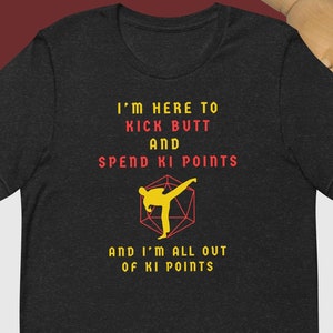 May include: A dark heather grey t-shirt with a yellow silhouette of a person kicking in a martial arts pose. The text "I'm here to kick butt and spend ki points and I'm all out of ki points" is printed in red and yellow on the shirt.