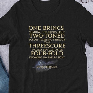 Puede incluir: Camiseta negra con un estampado gráfico dorado que dice "ONE BRINGS SHADOW, ONE BRINGS LIGHT TWO-TONED ECHOES TUMBLING THROUGH TIME THREESCORE WASTED, TEN CAST ASIDE FOUR-FOLD KNOWING, NO END IN SIGHT" y el texto "SHADOWBRINGERS" en la parte inferior.