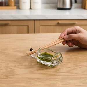 Op de afbeelding: Een helder glazen katvormig bakje met een groene vloeistof, met een paar houten eetstokjes erop. Het bakje staat op een lichtgekleurd houten oppervlak. Een hand houdt de eetstokjes vast.