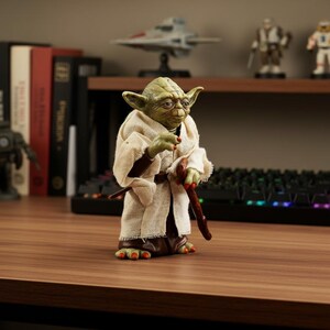 May include: A green-skinned humanoid figure with large pointed ears, wearing a textured beige robe over a dark brown garment. The figure has three-fingered hands and feet with orange-tipped digits, and is holding a dark brown wooden walking stick.