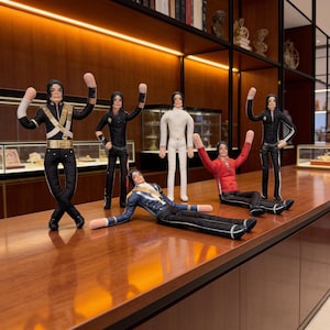 May include: A collection of five fabric dolls depicting a performer in various iconic outfits, including a black military-style jacket with gold accents, a white suit, a red jacket, and black trousers, all featuring long arms and detailed facial features.