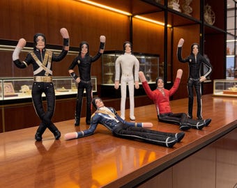 May include: A collection of five fabric dolls depicting a performer in various iconic outfits, including a black military-style jacket with gold accents, a white suit, a red jacket, and black trousers, all featuring long arms and detailed facial features.