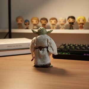 May include: A small green figurine of a character with pointed ears, wearing a textured, off-white fabric robe with a brown belt. The figure is viewed from the back, showing the frayed hem of the garment and the detailed folds of the fabric.