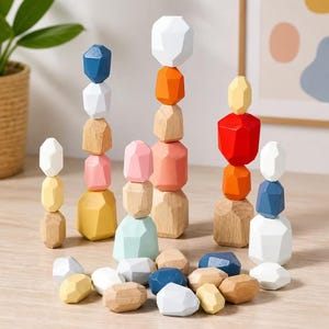 Puede incluir: Piedras apilables de madera en varios colores, incluyendo blanco, azul, naranja, rosa y amarillo. Las piedras están dispuestas en torres y dispersas sobre una superficie marrón claro. Las formas geométricas están diseñadas para jugar y equilibrar.