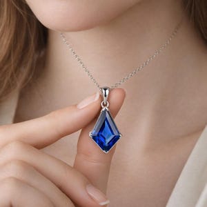Può includere: Una collana in argento con un ciondolo a forma di aquilone che presenta una gemma blu profondo sfaccettata. Il ciondolo è fissato tramite una contromaglia in argento a una sottile catenina, con un design geometrico dai bordi netti.