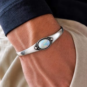 Op de afbeelding: Een zilveren armband met een centrale ovaalvormige iriserende opaalsteen. De steen is omlijst door een gedraaide metalen rand in touwstijl en geaccentueerd met twee kleine zilveren bolletjes aan weerszijden op een gladde, gepolijste zilveren band.