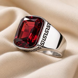 May include: A silver-toned ring featuring a large, rectangular, faceted deep red gemstone set in the centre. The band of the ring is wide and polished, decorated with a black engraved Greek key pattern that wraps around the sides of the setting.
