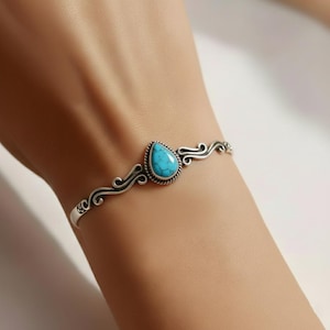 Peut inclure: Un bracelet manchette en argent orné d'une pierre turquoise en forme de goutte au centre, présentant des veines sombres. La bande métallique est décorée de motifs de volutes complexes de chaque côté de la pierre, offrant un aspect détaillé.