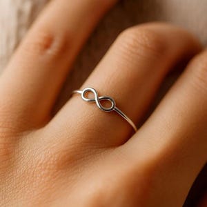 Puede incluir: Un anillo fino de tono plateado pulido con un símbolo de infinito en el centro. La banda es delgada y circular, descansando sobre un dedo, mostrando un acabado metálico liso y continuo que forma la figura horizontal del ocho.
