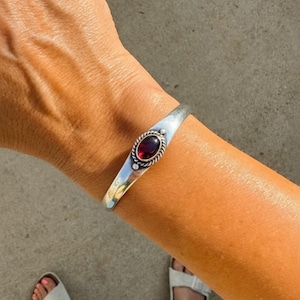 Pode incluir: Uma pulseira de prata polida com uma pedra preciosa granada vermelho-escura de formato oval no centro. A pedra está envolvida por uma borda de metal em estilo de corda trançada com pequenos detalhes de contas de prata no topo e na base.