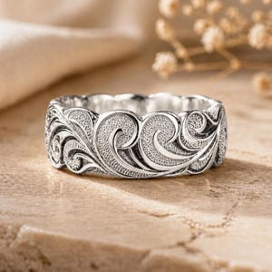 May include: A silver ring featuring a continuous band with an intricate, raised scrollwork pattern. The design consists of swirling, wave-like motifs with a textured, matte finish within the recessed areas, contrasting against the polished silver edges.