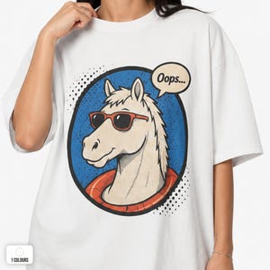 May include: A white t-shirt featuring a graphic print of a light-colored horse wearing red sunglasses and an orange life preserver. The horse is inside a blue circular frame with black dots, and a speech bubble above the horse contains the text Oops...
