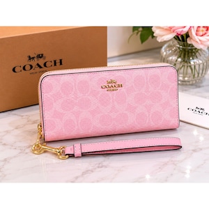 May include: A light pink zip-around wallet featuring a tonal signature C pattern, gold-tone hardware, and a detachable wrist strap. The front displays a gold metallic Coach logo. The wallet is accompanied by a branded box and a small white card.