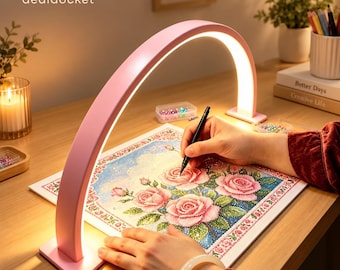 May include: A pink arched LED desk lamp illuminates a square diamond painting canvas featuring a rose floral design. The lamp has a curved frame with a light strip on the underside. The canvas displays pink roses and green leaves.