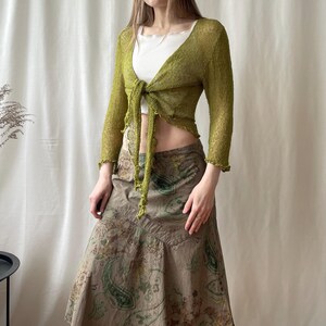 May include: A long-sleeved olive green tie-front cardigan with a textured finish, worn over a white ribbed tank top. The outfit is paired with a long, brown, A-line skirt featuring a dark green and tan paisley print pattern throughout the fabric.