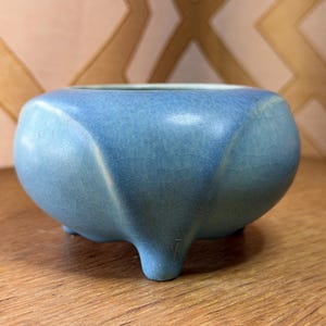 May include: A small, light blue ceramic bowl with a rounded shape and three small feet. The bowl has a textured surface and a crackled glaze. The interior is a slightly darker shade of blue. The bowl is sitting on a wooden surface.