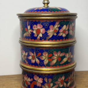 May include: A three-tiered, round trinket box with a lid. The box features a blue background with pink and red floral designs. The edges are trimmed with gold-colored metal. The lid is topped with a gold-colored conical finial.