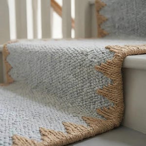 May include: A light grey stair runner with a textured, woven surface and a thick, tan jute border featuring a repeating triangular sawtooth pattern along the edges. The runner covers the horizontal treads and vertical risers of the staircase.