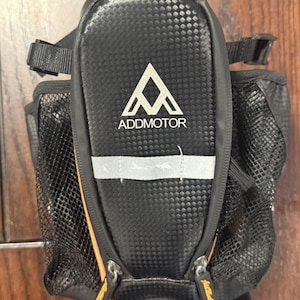 May include: A black bicycle saddle bag with a carbon fiber texture, featuring the white Addmotor logo and a horizontal reflective strip. The bag includes side mesh pockets, two zipper pulls with branding, and adjustable black attachment straps.