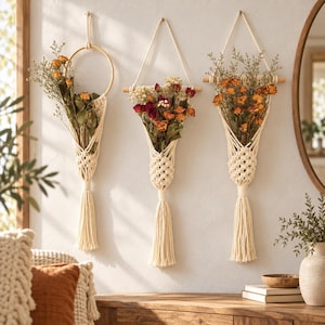 May include: Three wall-hanging macrame plant holders made of off-white cord, each containing a bouquet of dried flowers in shades of orange, red, and yellow. The holders feature intricate knotted patterns, wooden supports, and long decorative tassels.