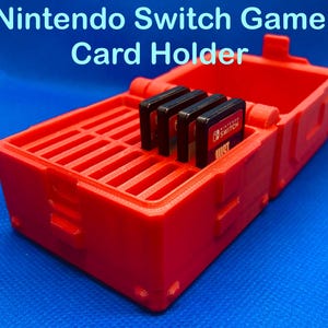 Può includere: Un porta schede di gioco per Nintendo Switch rettangolare di colore rosso brillante con coperchio incernierato. L'interno presenta una griglia di slot per organizzare le cartucce, con quattro cartucce nere per Nintendo Switch inserite.