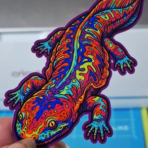 May include: A die-cut sticker featuring a stylized salamander with a vibrant, psychedelic color palette of neon orange, bright blue, lime green, and deep purple. The creature is depicted in a top-down view with intricate, swirling patterns.