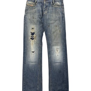 May include: Blue denim jeans featuring a distressed finish with frayed rips on the right leg, faded areas on the knees, and subtle brown staining. The jeans include a button fly, belt loops, and a small branded logo tag on the front pocket.