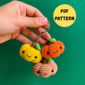 May include: A crochet keychain featuring three small, stuffed pumpkins in yellow, orange, and light brown. Each pumpkin has a dark brown stem, a green leaf, black bead eyes, and a stitched smile. A yellow circular graphic reads PDF PATTERN.