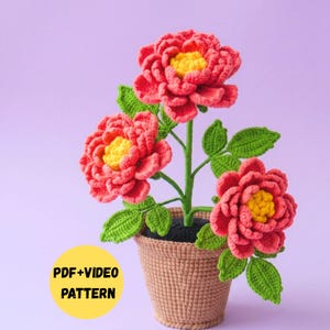 May include: A crochet flower arrangement featuring three vibrant pink peonies with bright yellow centers, green leaves, and stems, potted in a textured tan container. A yellow circular graphic displays the text PDF+VIDEO PATTERN.
