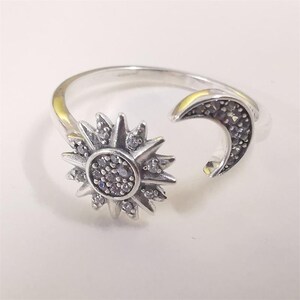 May include: A silver-toned open ring featuring a sun motif on one end and a crescent moon motif on the other. Both celestial shapes are embellished with small, clear, sparkling stones set into the metal surface, creating a detailed design.