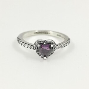May include: A silver ring featuring a central heart-shaped purple stone surrounded by a halo of small clear stones. The band is also set with a row of small clear stones along each side. The interior of the band is engraved with PANDORA and S925 ALE.