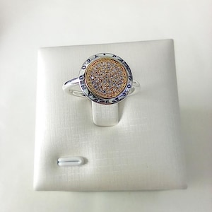 May include: A silver ring featuring a circular face with a central gold-toned disc encrusted with numerous small clear stones. The outer silver border of the circular face is engraved with the text PANDORA, separated by small dots.