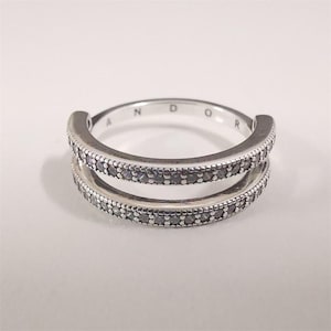 May include: A silver-toned double-band ring featuring two parallel rows of small, dark, circular stones set into the metal. The inner surface of the band is engraved with the partial text PANDOR, visible along the upper curve of the ring.