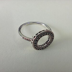 May include: A silver-coloured ring featuring a circular centrepiece adorned with numerous small, clear stones. The outer edge of the circular setting displays a repeating heart-shaped cutout pattern, and the band is also embellished with small stones.