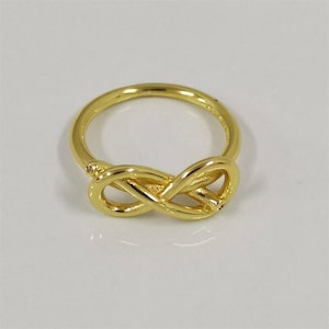May include: A polished gold-tone ring featuring a central infinity symbol design. The band is smooth and circular, connecting to the continuous looped figure-eight shape that sits prominently at the front of the jewelry piece.
