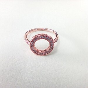 May include: A rose gold ring featuring a circular centrepiece adorned with small, clear pavé-set stones. The band is thin and metallic, with the circular design element centred on the top, showcasing a consistent rose gold finish throughout.