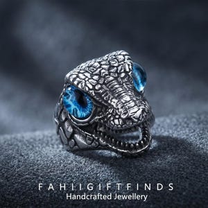 May include: A silver-colored ring shaped like a dinosaur head with textured scales and bright blue eyes. The ring has an open mouth and the text "FAHII GIFTFINDS Handcrafted Jewellery" at the bottom.